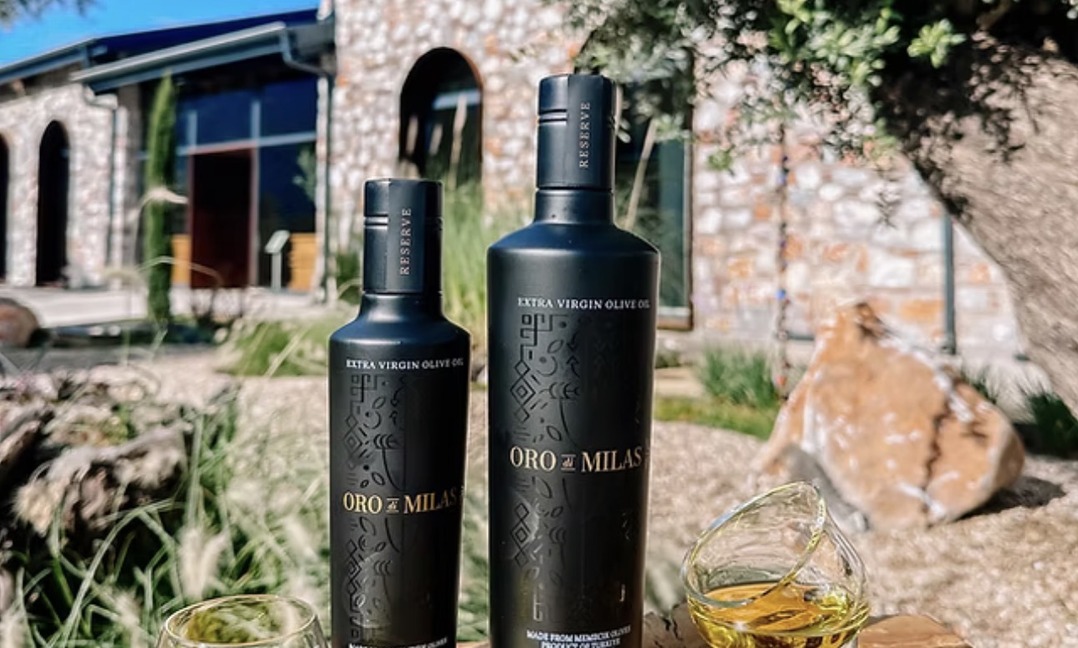 Zwei mattschwarze Oro di Milas Reserve Flaschen mit goldenem Olivenöl im Verkostungsglas vor einem historischen Steinhaus im Olivenhain